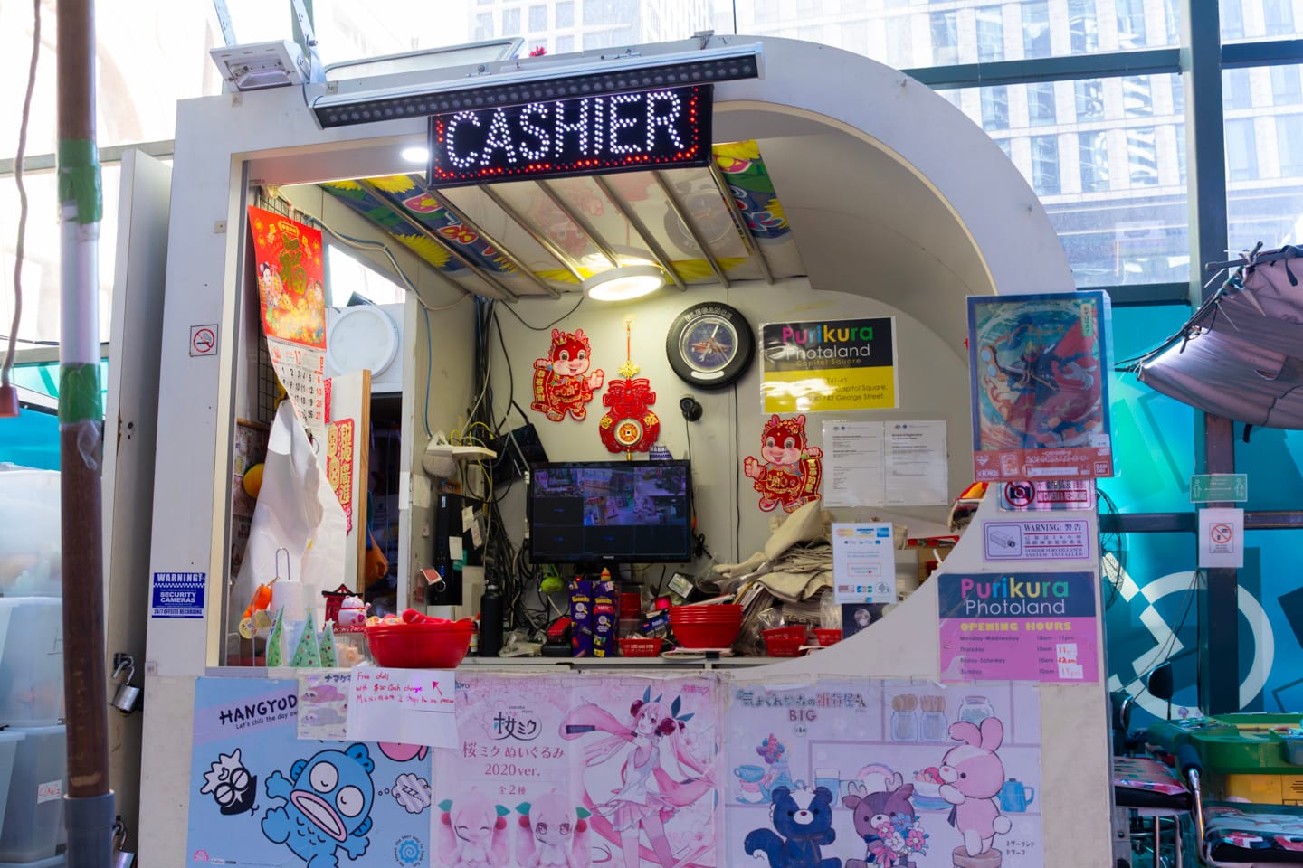 Purikura Photoland’s cashier booth. Various promotional posters, trading hours, Chinese calendars and decorations, CCTV monitors, and a clock, amongst other things, are affixed to the inside and outside of the walls.