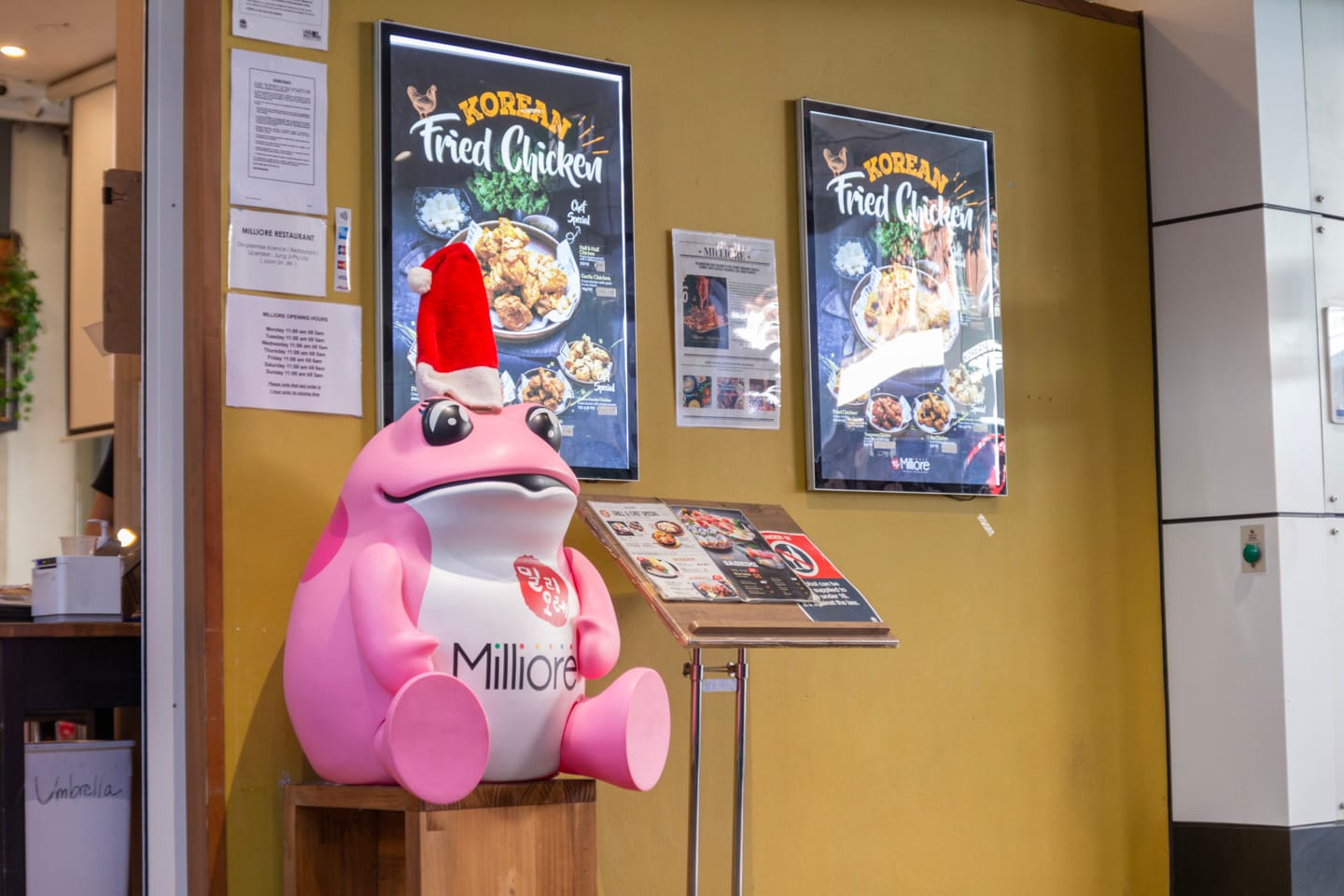A pink cartoonish frog sculpture with a Santa hat on its head, sitting on a wooden chair next to the menu of Milliore Restaurant.