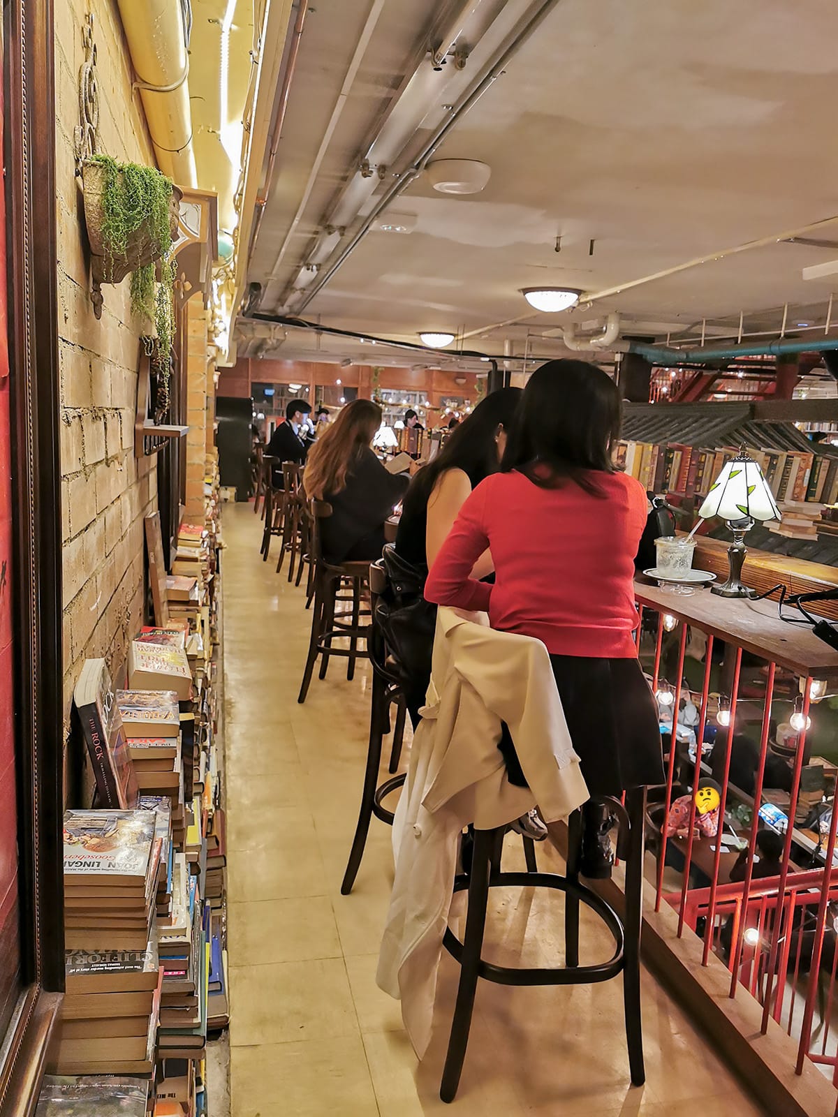 The upper floor of the left walkway just after going up the stairs. Paperback books line the edge of the wall, whilst visitors sit on high stools on the other side.